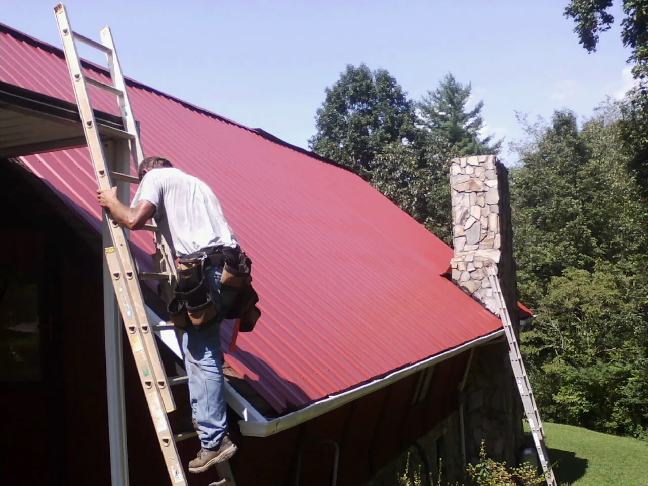 Roofer on a ladder beside a red metal roof and stone chimney — Soffit & Fascia Repair by Roofing Brothers in Marcellus
